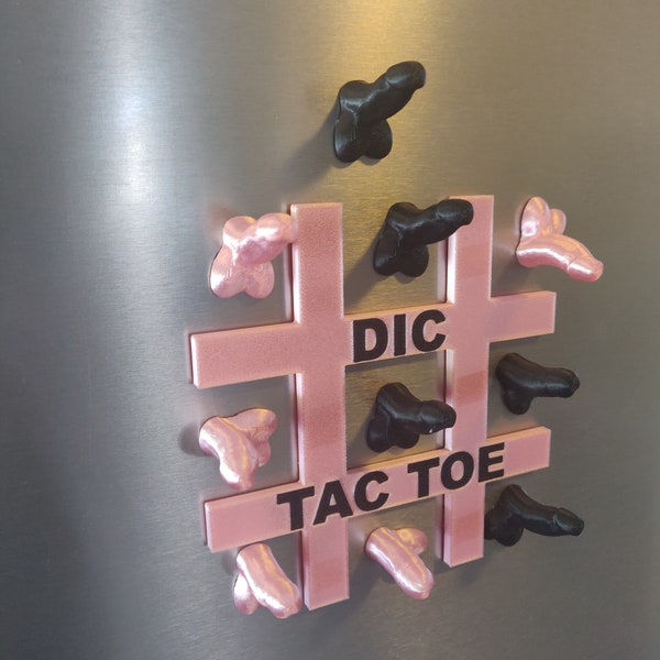 May include: A novelty Tic-Tac-Toe game with a pink grid and black and pink phallic-shaped game pieces. The words "DIC TAC TOE" are printed on the grid. The game is displayed on a metallic surface.