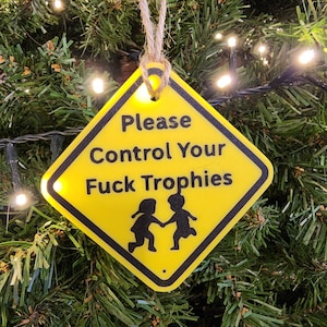 May include: A yellow diamond-shaped Christmas tree decoration with black text reading "Please Control Your Fuck Trophies." The ornament shows a black silhouette of two children holding hands, suspended by a twine string.