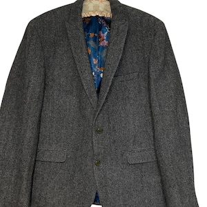 May include: A grey herringbone blazer with a two-button fastening and notched lapels. The blazer features two front pockets and a floral patterned lining in blue, pink, and orange. The blazer is suspended from a hanger.