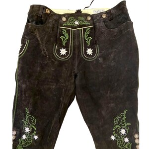 May include: Dark brown leather breeches featuring green embroidered vine patterns and white edelweiss flower motifs on the front pockets and lower legs. The garment includes a button-up front flap, belt loops, and metallic buttons.