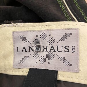 May include: A dark brown garment featuring a cream-colored interior label with the text LANHAUS C&A. The label is decorated with a black cross-stitch pattern and features a clear, round, four-hole button centered over the brand name.