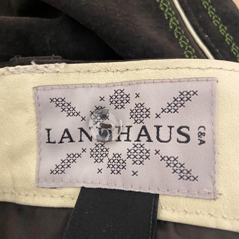 May include: A dark brown garment featuring a cream-colored interior label with the text LANHAUS C&A. The label is decorated with a black cross-stitch pattern and features a clear, round, four-hole button centered over the brand name.