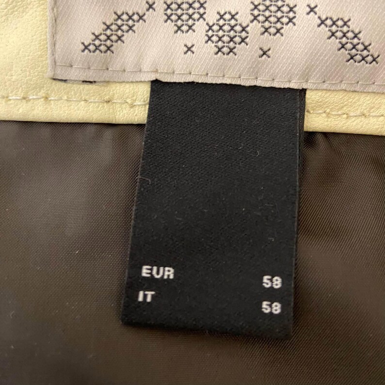 May include: A black clothing tag attached to a light yellow leather garment with a brown lining. The tag features the text EUR 58 and IT 58. Above the black tag is a light-colored fabric label with a dark gray cross-stitch pattern.