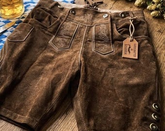 May include: Brown leather shorts with decorative stitching on the pockets and front. The shorts have a button closure and a small tag that says "Handcrafted in Bavaria". The shorts are likely part of a traditional German or Bavarian outfit.