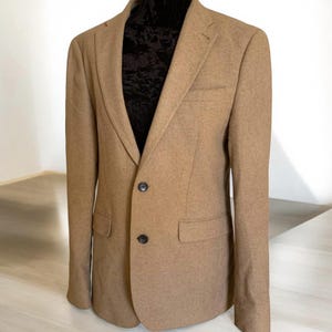 Può includere: Un blazer marrone con chiusura a due bottoni e una tasca singola sul lato sinistro.