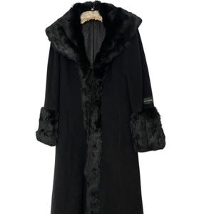 May include: A long black coat with a hood and fur trim. The coat features fur cuffs and trim along the front. A small label is visible on the sleeve. The coat is hanging on a wooden hanger.
