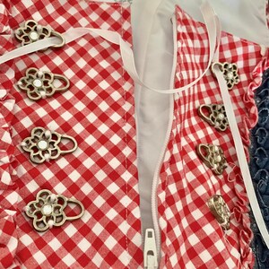 May include: Red and white gingham fabric with a zipper closure and decorative metal buttons. The fabric has a white ribbon tied around it.
