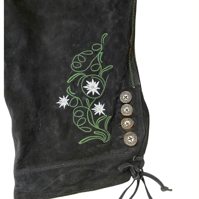 May include: Black suede trousers featuring green embroidered vine patterns and white edelweiss flower motifs. The side leg opening includes four round, textured metallic buttons and a thin leather lace tie at the bottom hem.
