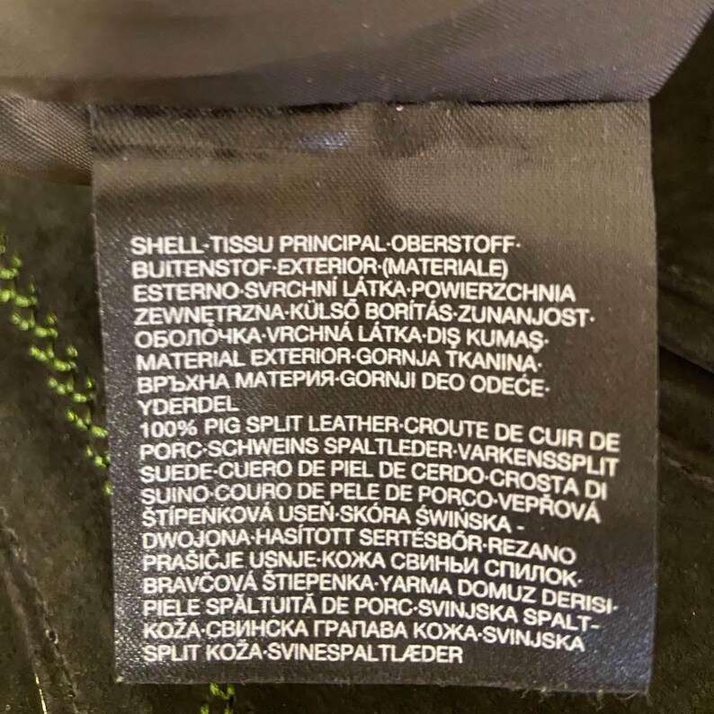 May include: A black fabric garment care label with white text. The label reads: SHELL-TISSU PRINCIPAL-OBERSTOFF-BUITENSTOF-EXTERIOR-(MATERIALE) ESTERNO-SVRCHN&Iacute; L&Aacute;TKA-POWIERZCHNIA ZEWNĘTRZNA-K&Uuml;LSŐ BOR&Iacute;T&Aacute;S-ZUNANJOST-ОБОЛОЧКА-VRCHN&Aacute; L&Aacute;TKA-DIŞ KUMAŞ-MATERIAL EXTERIOR-GORNJA TKANINA-ВРЪХНА МАТЕРИЯ-GORNJI DEO ODEĆE-YDERDEL 100% PIG SPLIT LEATHER-CROUTE DE CUIR DE PORC-SCHWEINS SPALTLEDER-VARKENSSPLIT SUEDE-CUERO DE PIEL DE CERDO-CROSTA DI SUINO-COURO DE PELE DE PORCO-VEPŘOV&Aacute; &Scaron;T&Iacute;PENKOV&Aacute; USEŇ-SK&Oacute;RA ŚWIŃSKA-DWOJONA-HAS&Iacute;TOTT SERT&Eacute;SBŐR-REZANO PRA&Scaron;IČJE USNJE-КОЖА СВИНЬИ СПИЛОК-BRAVČOV&Aacute; &Scaron;TIEPENKA-YARMA DOMUZ DERISI-PIELE SPĂLTUITĂ DE PORC-SVINJSKA SPALT-KOŽA-СВИНСКА ГРАПАВА КОЖА-SVINJSKA SPLIT KOŽA-SVINESPALTLAEDER.