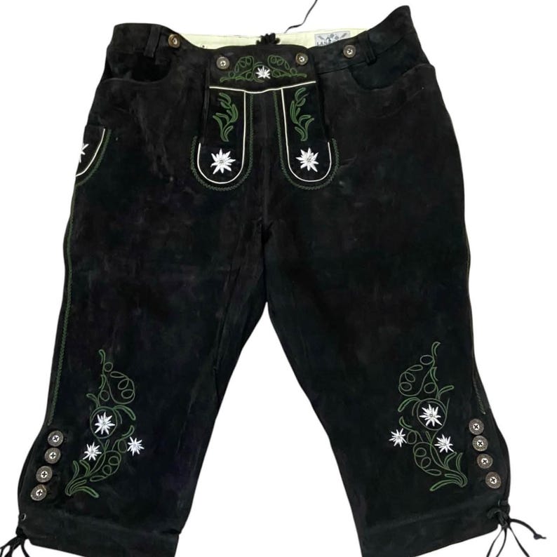 May include: Black leather knee-length breeches featuring green vine embroidery and white edelweiss flower motifs on the front flap and lower legs. The garment includes decorative metal buttons along the waist and the lower side cuffs.