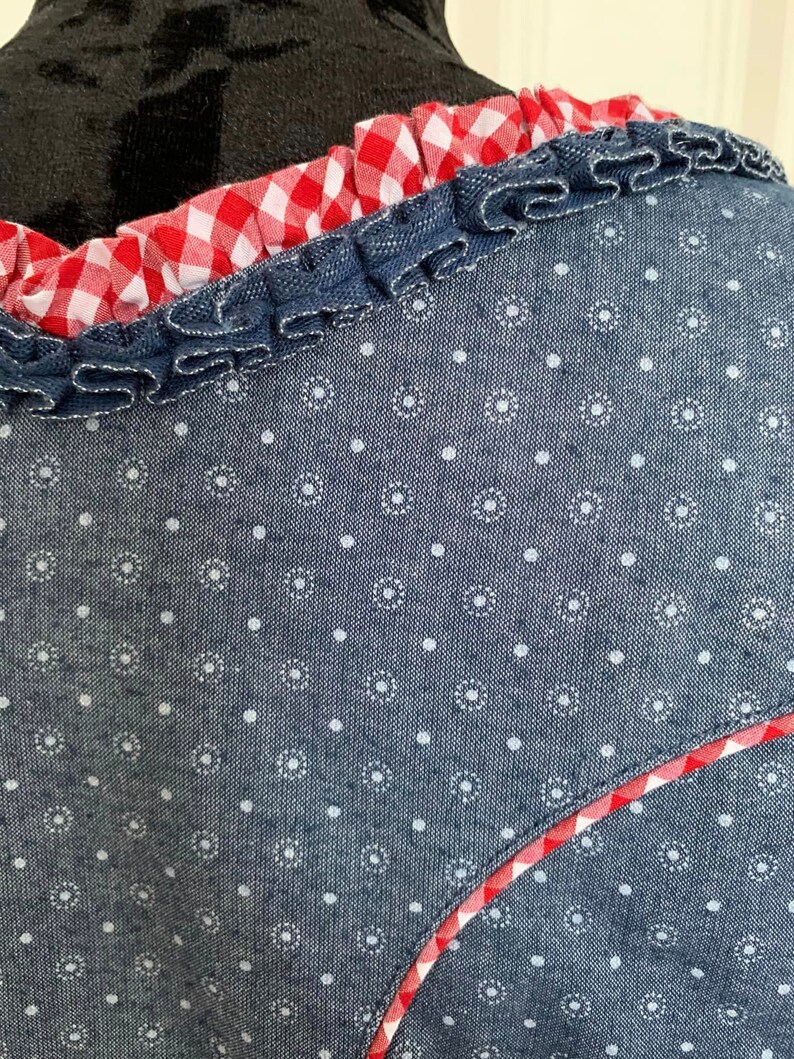 May include: A blue denim top with a white polka dot pattern and a red and white gingham trim. The trim is ruffled and sewn around the neckline.