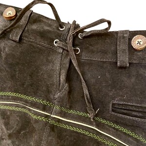 May include: Dark brown suede leather trousers featuring a lace-up back detail with thin leather cords, belt loops, and decorative light green embroidered stitching across the front. The waistband includes round, light-colored buttons.