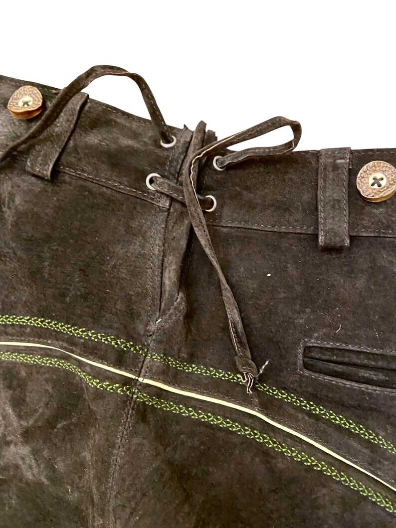 May include: Dark brown suede leather trousers featuring a lace-up back detail with thin leather cords, belt loops, and decorative light green embroidered stitching across the front. The waistband includes round, light-colored buttons.