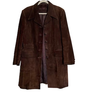 May include: A dark brown suede coat featuring a pointed collar, a front button closure, and two flap pockets on the sides. The interior label displays the text YESSICA. The garment has long sleeves and a structured, knee-length silhouette.