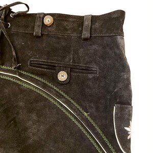 May include: A pair of black suede shorts featuring a high waistband with belt loops, a front pocket with a button closure, and decorative green braided trim. The shorts include light-colored piping and a small embroidered detail on the side.