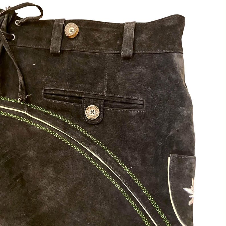 May include: A pair of black suede shorts featuring a high waistband with belt loops, a front pocket with a button closure, and decorative green braided trim. The shorts include light-colored piping and a small embroidered detail on the side.