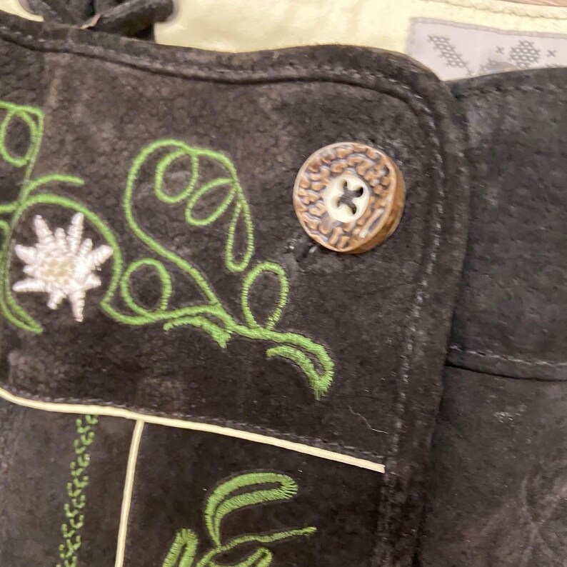 May include: A dark brown suede garment featuring green embroidered vine patterns and a white edelweiss flower motif. The piece includes a textured, round, light brown button with a four-hole center and light-colored piping along the seams.
