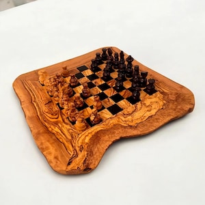 Op de afbeelding: Een houten schaakspel met een natuurlijke, organische rand. Het schaakbord heeft een zwart-bruin geblokt patroon, met licht- en donkerbruine schaakstukken. De set wordt gepresenteerd op een wit oppervlak.