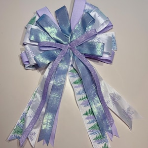 May include: A decorative bow made from layered ribbons in shades of blue, purple, and white. Some ribbons have a shimmering finish, while others feature a winter forest and snowflake pattern. The bow is tied in the centre.