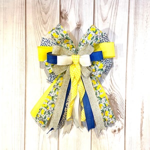 May include: A decorative bow with a lemon theme. The bow is made from ribbons in yellow, blue, white, and patterned fabrics with lemon and floral designs. It is attached to a light-coloured wooden surface.