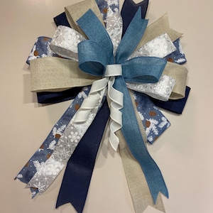 May include: A blue, white and beige bow with a winter theme. The bow is made of various ribbons with different patterns, including snowflakes and pine cones.