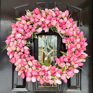 May include: A vibrant pink tulip wreath. The wreath is densely packed with various shades of pink tulips, creating a full and lush appearance. Green leaves are interspersed throughout the design. The wreath is displayed on a black door.
