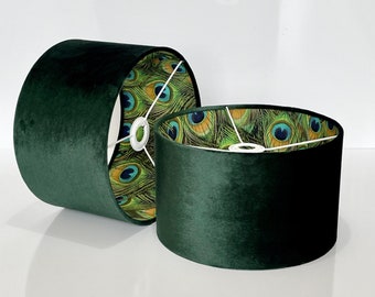 May include: Two dark green velvet lampshades. The interior of the lampshades features a pattern of peacock feathers in shades of green, blue, and gold. The lampshades are cylindrical and have a white metal frame.
