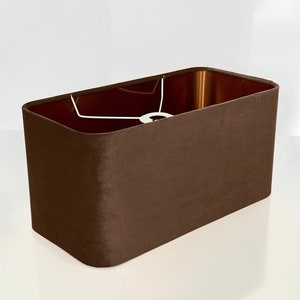 May include: A rectangular lampshade in a rich, dark brown color. The shade has a smooth, velvety texture and a copper-colored interior. A white metal frame is visible inside the shade, designed to hold a light bulb. The lampshade is a home decor item.