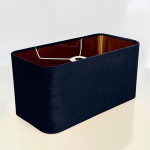 Op de afbeelding: Een rechthoekige lampenkap van donkerblauw fluweel. De kap heeft een witte binnenkant en een koperkleurige voering.
