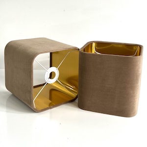 Könnte beinhalten: Zwei quadratische Lampenschirme mit goldenem Innenleben. Die Lampenschirme sind aus braunem Stoff mit weicher Textur.