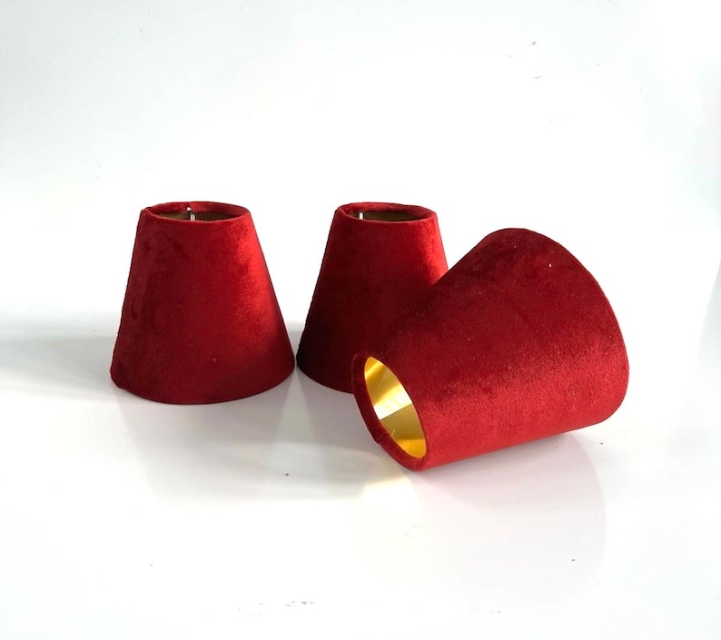May include: Three red velvet lampshades with gold trim. The lampshades are cone-shaped and have a smooth, soft texture.