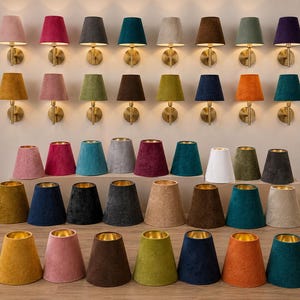 May include: A collection of wall-mounted lamps with brass fixtures and fabric shades in various colours, alongside a display of matching conical fabric lampshades with gold-toned interiors arranged in rows on a flat surface.