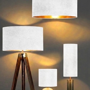 Può includere: Una collezione di lampade moderne con paralumi bianchi. Le lampade includono una lampada a sospensione, una lampada da terra con base a treppiede in legno e due lampade da tavolo. La lampada a sospensione ha l'interno dorato. Le lampade sono illuminate.