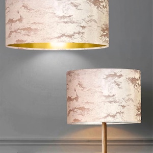 May include: A cylindrical pendant lamp shade and a matching table lamp shade, both featuring a textured, mottled cream and rose gold finish. The pendant shade has a reflective gold interior, while the table lamp sits on a light wood tripod base.