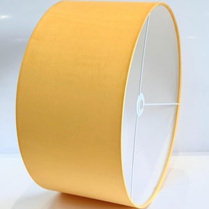 May include: A cylindrical lampshade in a vibrant yellow hue. The lampshade has a soft, velvety texture and a white interior. The metal frame is visible inside the shade. This lampshade is designed for use with a lamp base.