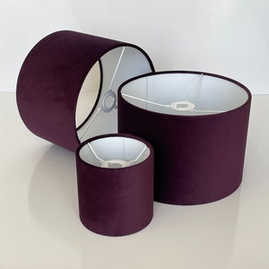 May include: Three purple velvet lampshades of different sizes. The lampshades are cylindrical and have a white interior.