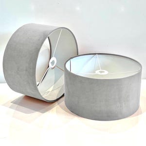 May include: Two gray lampshades with white fabric interiors. The lampshades are cylindrical and have a smooth, velvety texture.