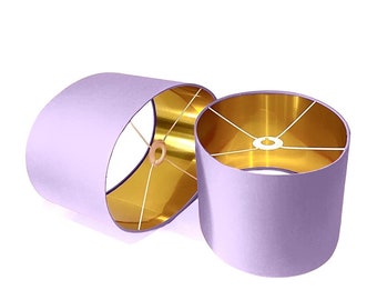 May include: Two oval-shaped lampshades with a lilac fabric exterior and a gold interior. The lampshades are sitting on a white surface.