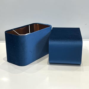 Può includere: Due paralumi in velluto blu. Uno è rettangolare con interno color rame e l'altro ha una forma quadrata curva più piccola. I paralumi sono di un blu intenso e ricco e sembrano essere fatti di un materiale morbido e soffice. Sono probabilmente per una lampada da tavolo o da terra.