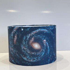 Peut inclure: Un abat-jour bleu et blanc avec un imprimé de galaxie. L'imprimé présente une nébuleuse tourbillonnante avec des étoiles et un centre orange vif.