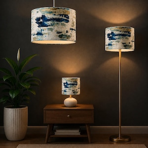 May include: A set of three lighting fixtures including a hanging pendant lamp, a table lamp with a ribbed white base, and a floor lamp with a thin gold stand. Each lamp features a cylindrical shade with a blue, gold, and white abstract paint pattern.