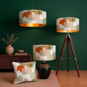 Könnte beinhalten: Eine Kollektion von Wohnaccessoires mit einem geometrischen Muster in Orange, Salbeigrün und Creme. Das Set besteht aus einer zylindrischen Hängeleuchte, einer Stativ-Stehleuchte, einer Tischleuchte mit schwarzem Fuß und einem quadratischen Kissen.