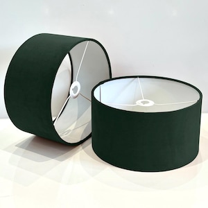 May include: Two dark green drum lampshades with white fabric interiors. The lampshades are made of a soft, velvety material.