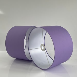 Pode incluir: Duas abajures cilíndricos em um tom roxo suave. As abajures têm um acabamento liso e fosco e um interior branco. A estrutura metálica é visível dentro das abajures. As abajures são projetadas para uso com uma base de lâmpada.