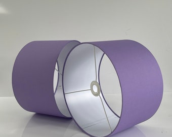 May include: Two cylindrical lampshades in a soft purple hue. The shades have a smooth, matte finish and a white interior. The metal frame is visible inside the shades. The lampshades are designed for use with a lamp base.