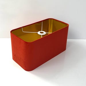 Könnte beinhalten: Rechteckiger Lampenschirm mit leuchtend roter Außenseite und goldfarbenem Innenraum. Der Schirm hat eine weiche, samtige Textur und ein weißes Metallgestell. Für eine Lampenbasis konzipiert.