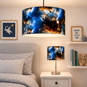 May include: A pendant light and a table lamp feature a celestial nebula design with shades of blue, orange, and white. The lamps are cylindrical and have a metal base. A framed astronaut art piece and a sign that says "Dream Big Little One" are also in the room.