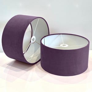 Può includere: Due paralumi in velluto viola con interni in tessuto bianco. I paralumi sono cilindrici e hanno un design semplice.