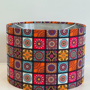 Peut inclure: Abat-jour cylindrique avec un motif patchwork vibrant. Le motif présente des carrés colorés avec des motifs floraux et géométriques complexes dans des tons d'orange, de rose, de bleu et de violet. L'intérieur est argenté, et l'esthétique générale est bohème.