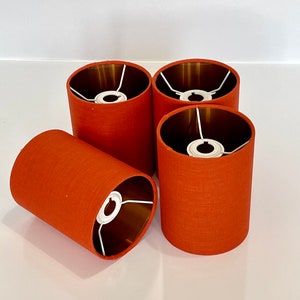 May include: Four orange fabric lampshades with gold interiors. The lampshades are cylindrical and have a white metal frame.